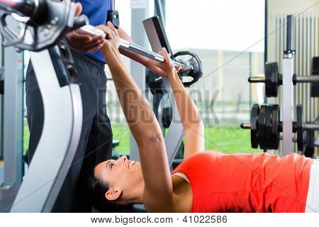 Picture or Photo of Woman with her personal fitness trainer in the gym exercising with dumbbells, she is using barbell on a weight bench