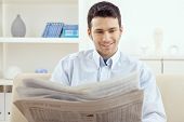 picture of newspaper  - Happy young man sitting on couch reading newspaper at home - JPG 