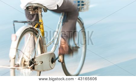 Picture or Photo of Focus on old white bicycle from behind. Motion blur of person riding bike outside. Popular transport in Vietnam, Asia. Legs of man on bicycle on blue background. Road travel, healthy lifestyle.