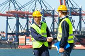 foto of industrial  - Two dockers shake hands in front of an industrial harbor with cranes and a container ship being unloaded - JPG 