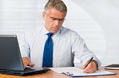 image of businessmen  - Mature businessman writing on paperwork and working with laptop in his office - JPG 