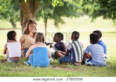Picture or Photo of Children and education young woman at work as educator reading book to boys and girls in park